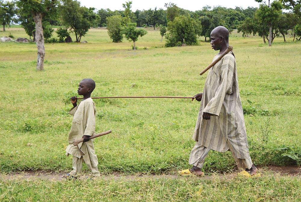 Masu, blinded by River Blindness, is guided by his young son along a dirt path in rural Nigeria, supported by cbm New Zealand